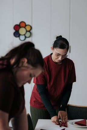 Zwei Frauen in roten Hemden falten Servietten an einem Tisch, eine fokussiert und eine leicht unscharf im Vordergrund.