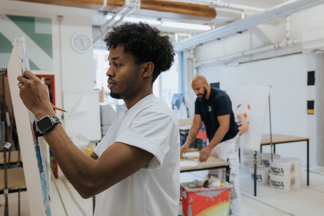 Zwei Männer, die in einem hellen Atelier an Kunstprojekten arbeiten; einer malt, während der andere an einem Tisch im Hintergrund arbeitet.