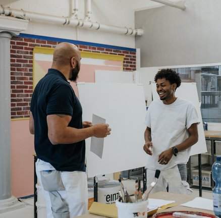 Zwei Männer unterhalten sich lächelnd in einem Kunstatelier, halten Papier in der Hand und stehen neben Malutensilien.