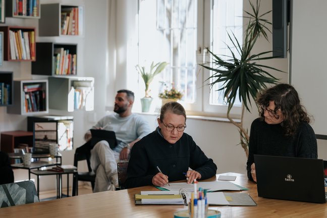 Zwei Frauen arbeiten an einem Tisch mit Büchern und einem Laptop; ein Mann sitzt lesend im Hintergrund.